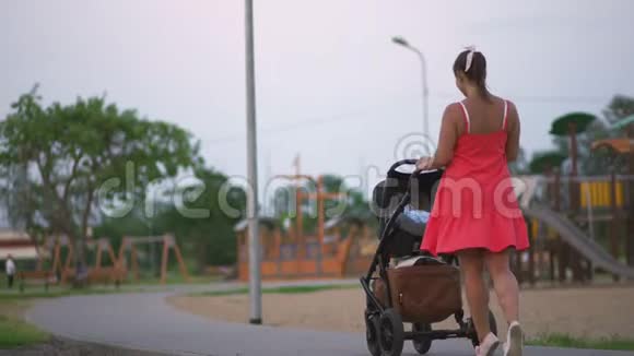 年轻的母亲走在城市公园的婴儿车上穿着鲜红的连衣裙赤着双腿视频的预览图