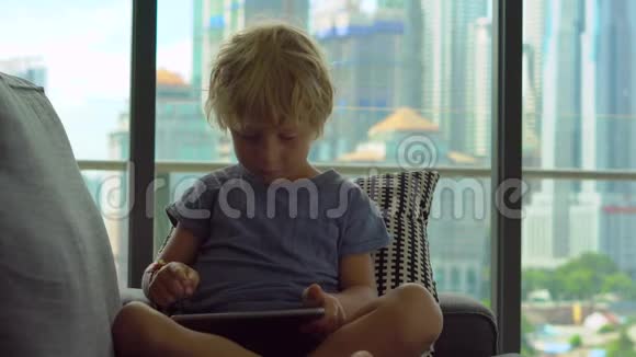 小男孩用平板电脑坐在一扇大窗户前背景是摩天大楼视频的预览图