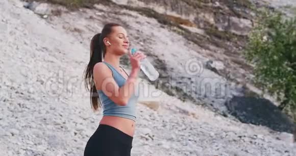 漂亮的运动型女士在进行了艰苦的锻炼后口渴地喝着水她列举了无线音乐视频的预览图