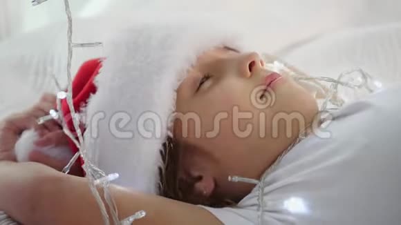 圣诞老人帽子里可爱的小宝宝的特写视频躺在床上玩挂在冷杉树枝上的花环视频的预览图