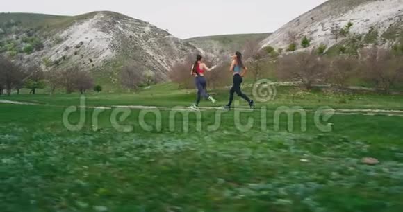 在一个完美的夏日里两个女人穿过一片绿色的田野和山景在大自然中锻炼身体视频的预览图
