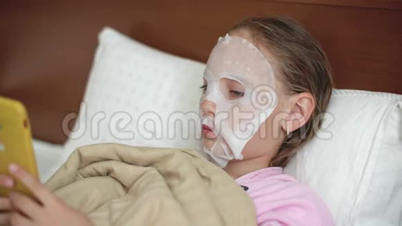 年轻女孩脸上戴着布面化妆品面膜躺坏了少女在化妆时使用智能手机视频的预览图