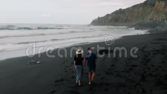 漂亮的电影镜头一对年轻夫妇在黑色的火山沙滩上散步然后用胳膊跑步鸟瞰图视频的预览图