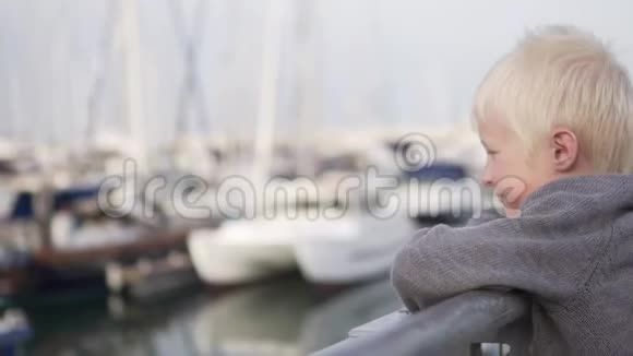一个悲伤的金发男孩站在游艇附近的码头上看着大海视频的预览图