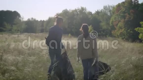 两个戴着手套拿着黑色塑料垃圾袋的女孩正走过田野志愿者收集垃圾视频的预览图