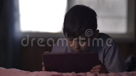 有趣的男孩躺在家里的床上用平板电脑玩得很开心视频的预览图