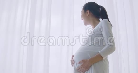 美丽的亚洲孕妇透过窗户望着客厅揉着肚子视频的预览图
