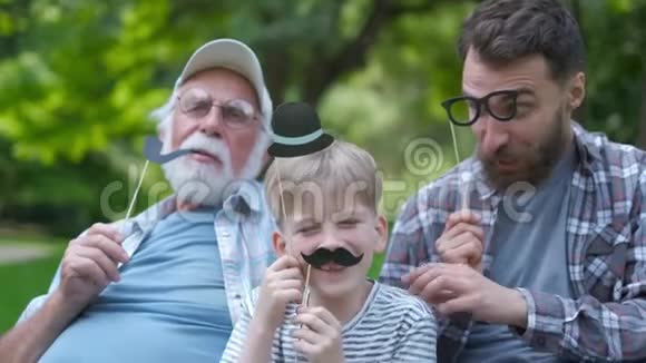 快乐有趣的家庭儿子和爸爸爷爷带着假胡子帽子眼镜在公园户外度假好日子视频的预览图