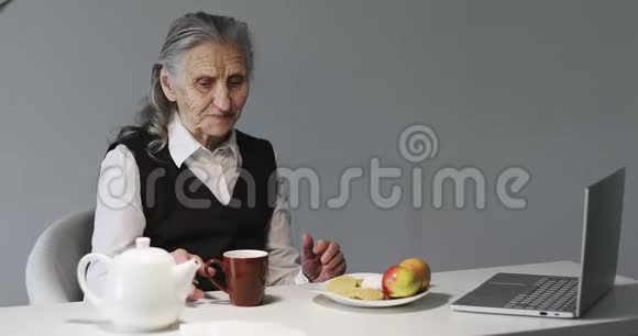 一位皱纹很深的老妇人在办公室吃早餐看着笔记本电脑视频的预览图