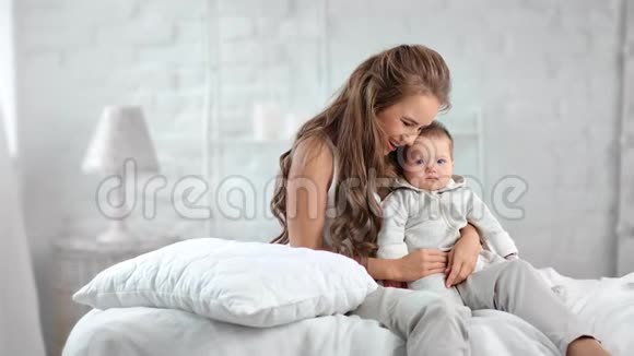 漂亮的女人放松可爱的婴儿坐在白色卧室的床上感觉到积极的情绪视频的预览图