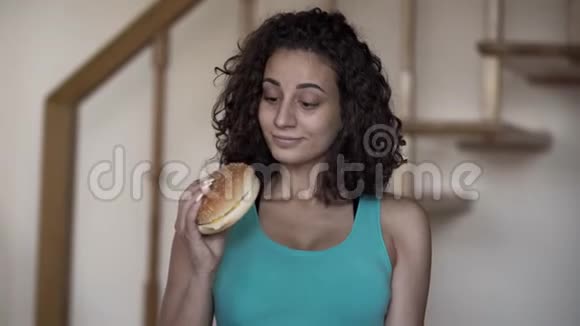 可爱的卷曲的年轻女人手里拿着一个大汉堡和一个小哑铃试图在一个健康的人之间做出选择的肖像视频的预览图
