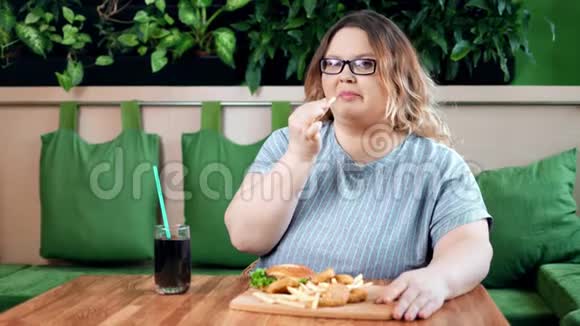 快乐的胖女人在咖啡馆里吃着开胃的炸薯条汉堡和金块看着镜头视频的预览图