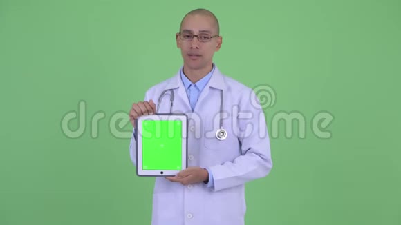 戴着帽子的秃头多民族医生一边说话一边显示数字平板电脑视频的预览图