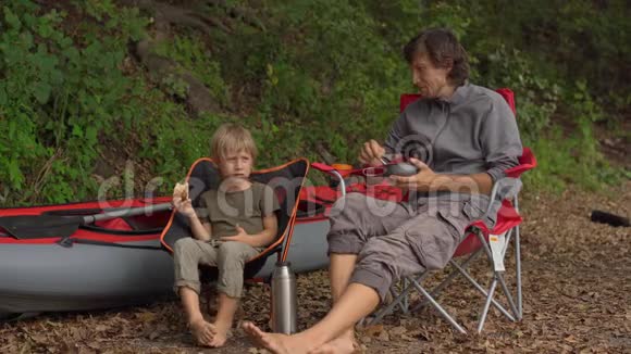 一个年轻人和他的小儿子在篝火旁休息他们坐在一个巨大的充气皮艇前视频的预览图