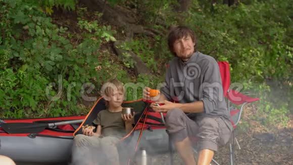 一个年轻人和他的小儿子在篝火旁休息他们坐在一个巨大的充气皮艇前视频的预览图