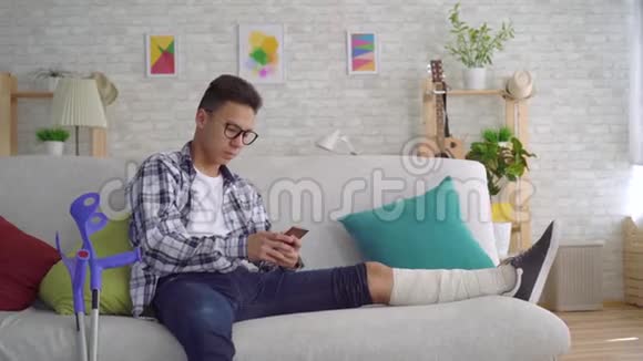 悲伤的亚洲年轻人一条绑着绷带的断腿躺在沙发上用智能手机看着摄像机视频的预览图