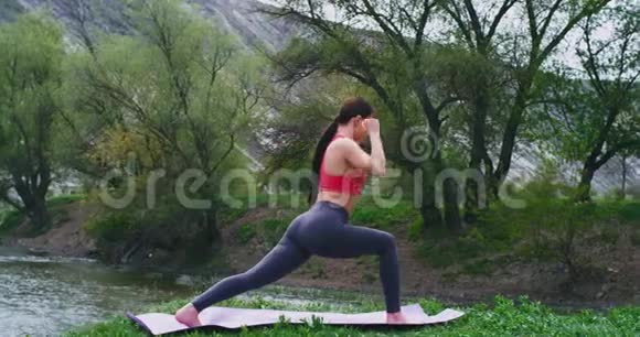 在岩石和河流景观中间年轻女士在垫子上练习瑜伽她做伸展身体的姿势视频的预览图