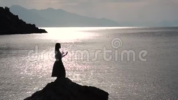 浪漫的女人在日出时在海边的岩石上缓慢地跳舞视频的预览图