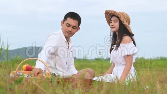 亚洲年轻夫妇在绿色草地野外背景下野餐男人把苹果给女人在调情蜜月之间吃饭视频的预览图
