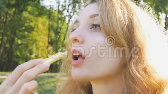 在公园里坐在长凳上吃炸薯条的年轻金发女人的户外肖像不健康饮食视频的预览图