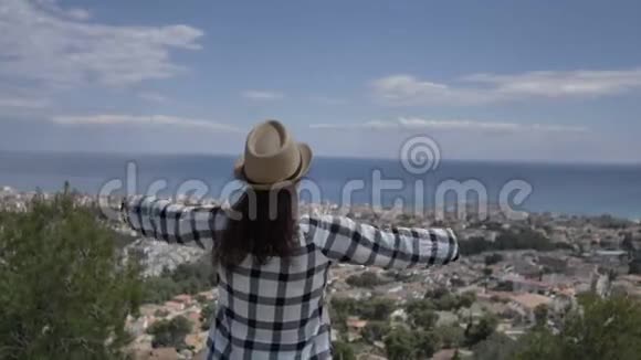 戴帽子的女孩站在靠近大海的顶端看着太阳举起双手飞向天空视频的预览图