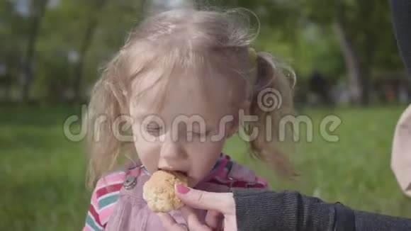 年轻母亲的手在公园里用一块饼干喂她可爱的金发女儿积极健康的生活方式视频的预览图