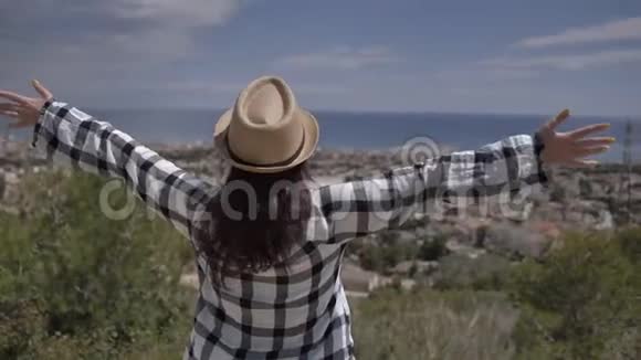 戴帽子的女孩站在靠近大海的顶端看着太阳举起双手飞向天空视频的预览图