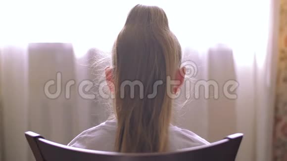 年轻的金发女孩头发尾巴坐在家里的前窗椅子上坐在椅子前窗前的女孩视频的预览图