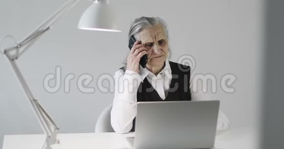 一位头发灰白皱纹深的严肃老妇人正在办公室的手机上聊天视频的预览图