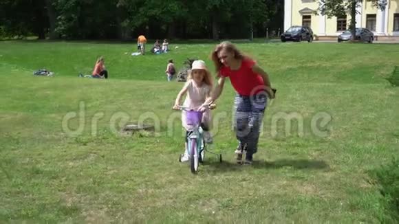 妈妈帮助她4岁的女儿骑自行车穿过公园草地视频的预览图