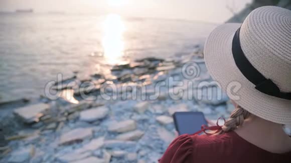 从后面的近景一个漂亮的年轻女孩穿着一件夏天的红色连衣裙和一顶草帽坐在海边的石头上视频的预览图