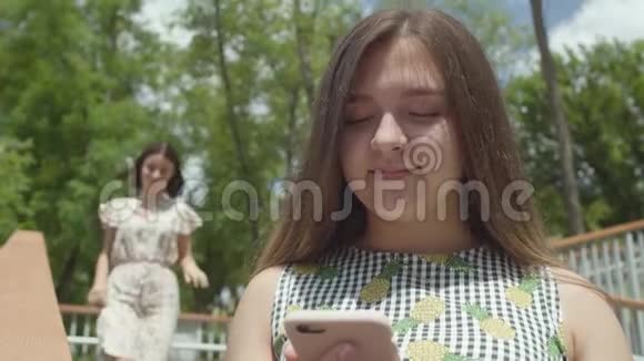 可爱的年轻女孩坐在前台在手机上发短信无忧无虑的朋友跑在她身后关上了视频的预览图