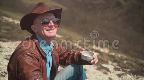 在山上一个男人戴着牛仔帽皮夹克蓝色牛仔裤和眼镜男人看着相框笑了笑视频的预览图