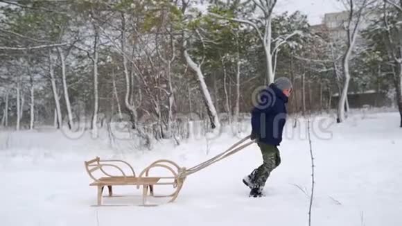 穿暖和衣服的可爱男孩在下雪的冬天拉木雪橇小男孩在公园里下雪跟踪视频的预览图