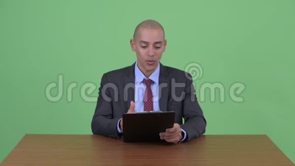 快乐秃顶的多民族商人在桌子后面展示剪贴板和竖起大拇指视频的预览图