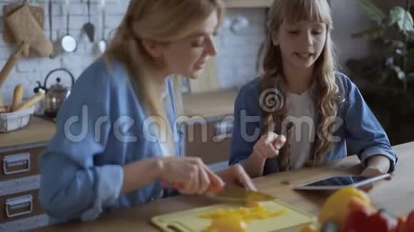 妈妈和女儿一起在一个明亮的大厨房做饭妈妈切多汁的辣椒一个女孩在平板电脑上读到一个食谱视频的预览图