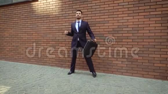 有魅力的男人留着胡子和公文包在城市街道上跳舞在现代砖墙的背景下视频的预览图