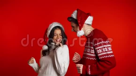 美丽的年轻夫妇戴着圣诞帽跳舞玩得开心在红工作室里欢乐视频的预览图