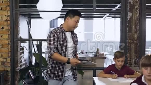 年轻的韩国小学男教师在课桌之间行走把电脑平板电脑放在课桌上供学生学习视频的预览图