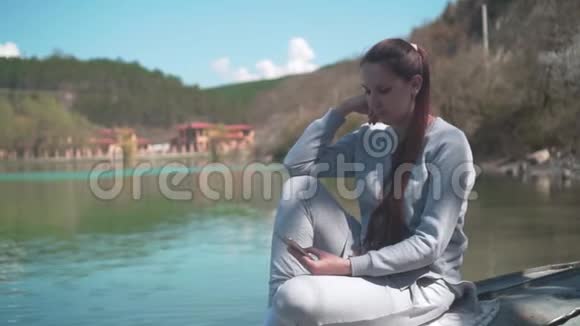 春天的一天一个年轻的女人坐在湖边的一艘旧船上看着电话在大自然中放松视频的预览图