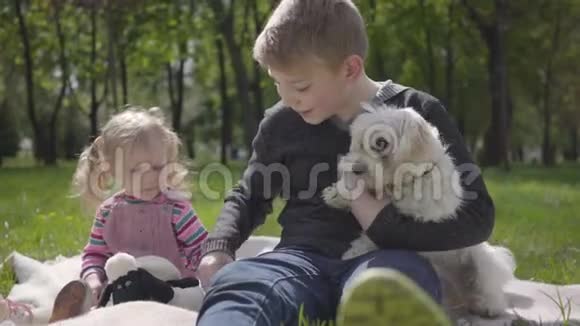 哥哥和妹妹坐在美丽的绿色公园的毯子里带着白色的毛茸茸的狗视频的预览图