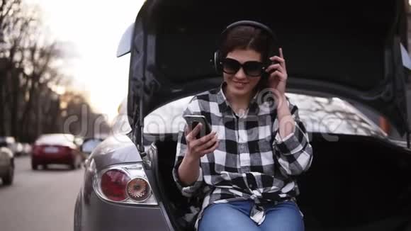 微笑的黑发女人坐在敞篷的车里在路的后备箱里听耳机里的音乐手机好的视频的预览图