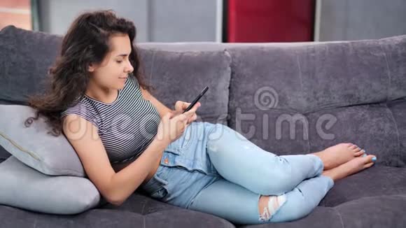 快乐轻松年轻居家女人躺在沙发上享受着智能手机聊天视频的预览图