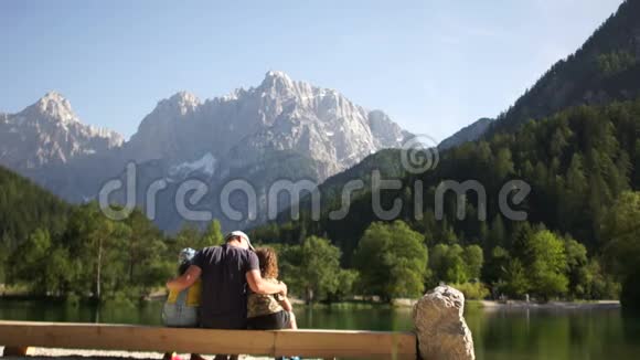 父亲和他的两个十几岁的孩子坐在一个山湖岸边的长凳上家人欣赏美丽的视频的预览图