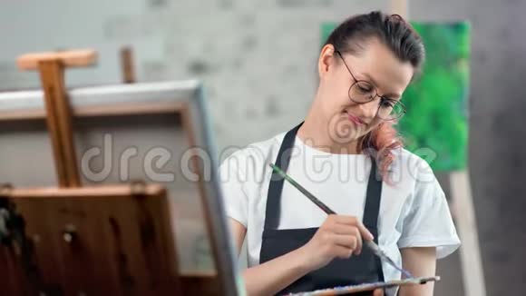 穿着围裙的年轻女子微笑着在艺术工作室中的特写镜头中欣赏绘画视频的预览图