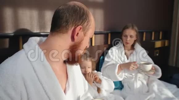 一家人穿着白色浴袍坐在酒店床上喝茶视频的预览图