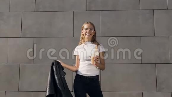一个年轻漂亮的女孩穿着白色的t恤和夹克在夏天的一天通过一根管子喝奶昔的肖像视频的预览图