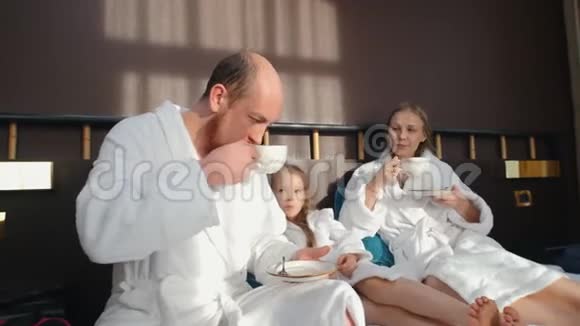 一家人穿着白色浴袍坐在床上喝茶视频的预览图