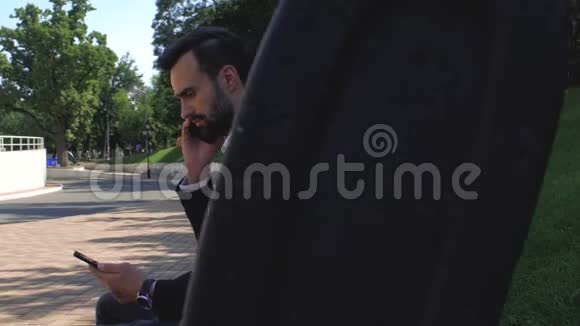 商人在长凳上的公园里用手机在手机上说话的人胡子男在智能手机上说话年轻青年视频的预览图