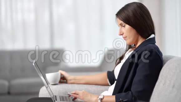自信的商务女性从杯子里喝咖啡用笔记本电脑在舒适的沙发上做中等特写视频的预览图